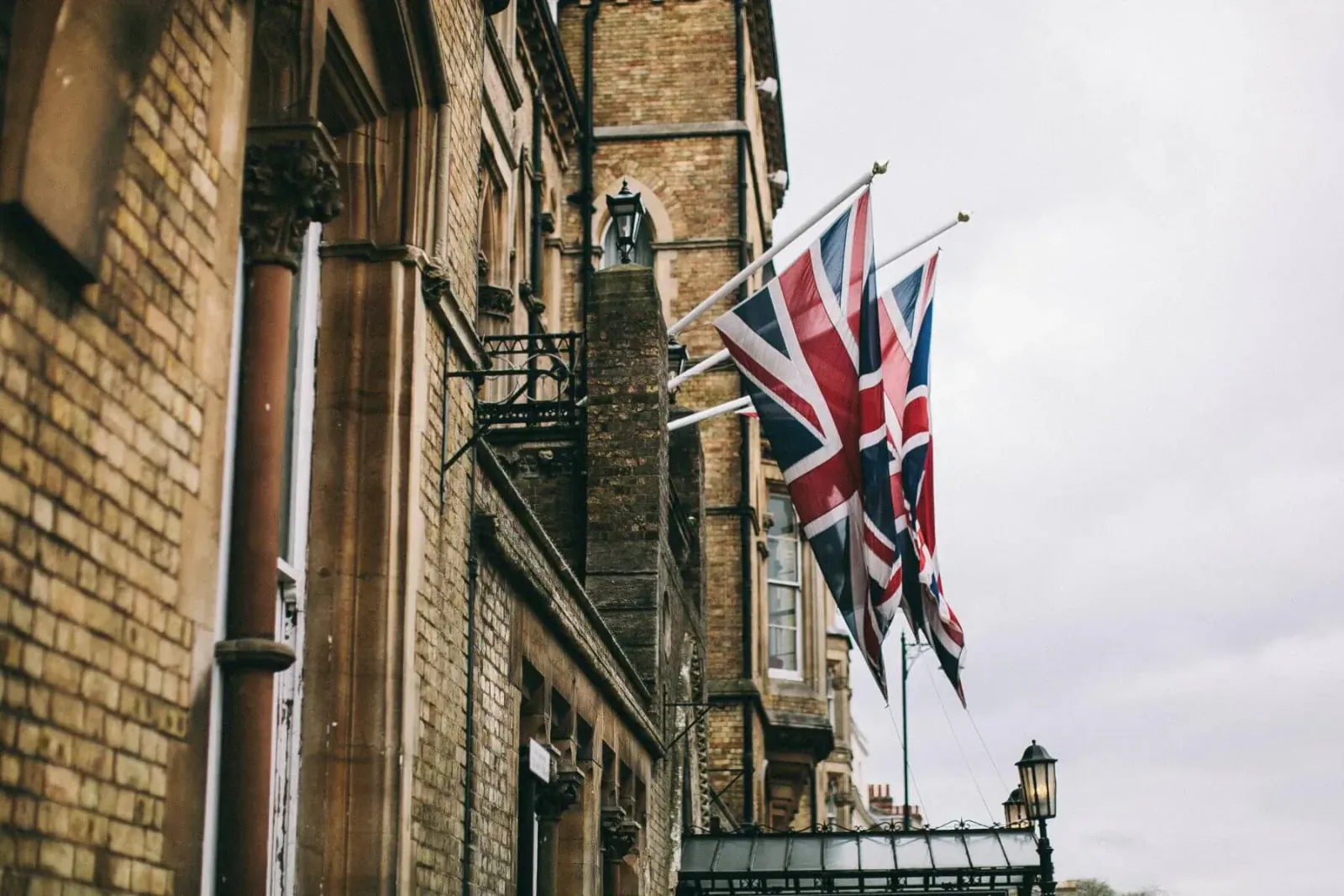 Three Union Jack flags waving against a historic brick building under cloudy skies, UK staycation vibe.