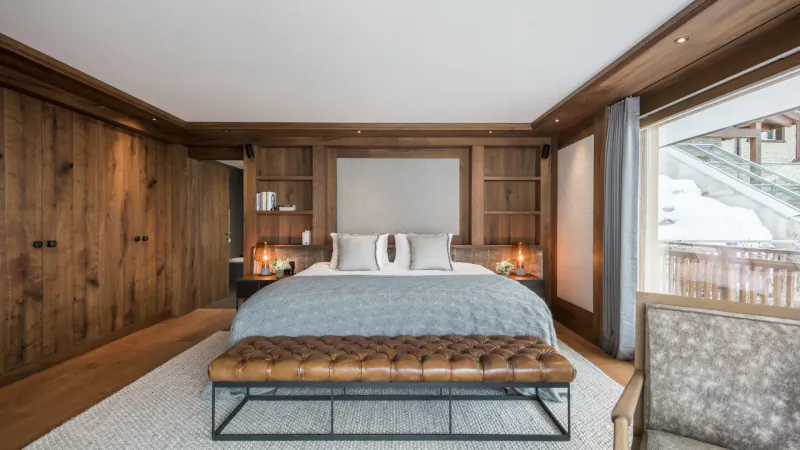 Luxury ski chalet bedroom with wooden walls, king bed, bench, armchair, and snowy mountain view.