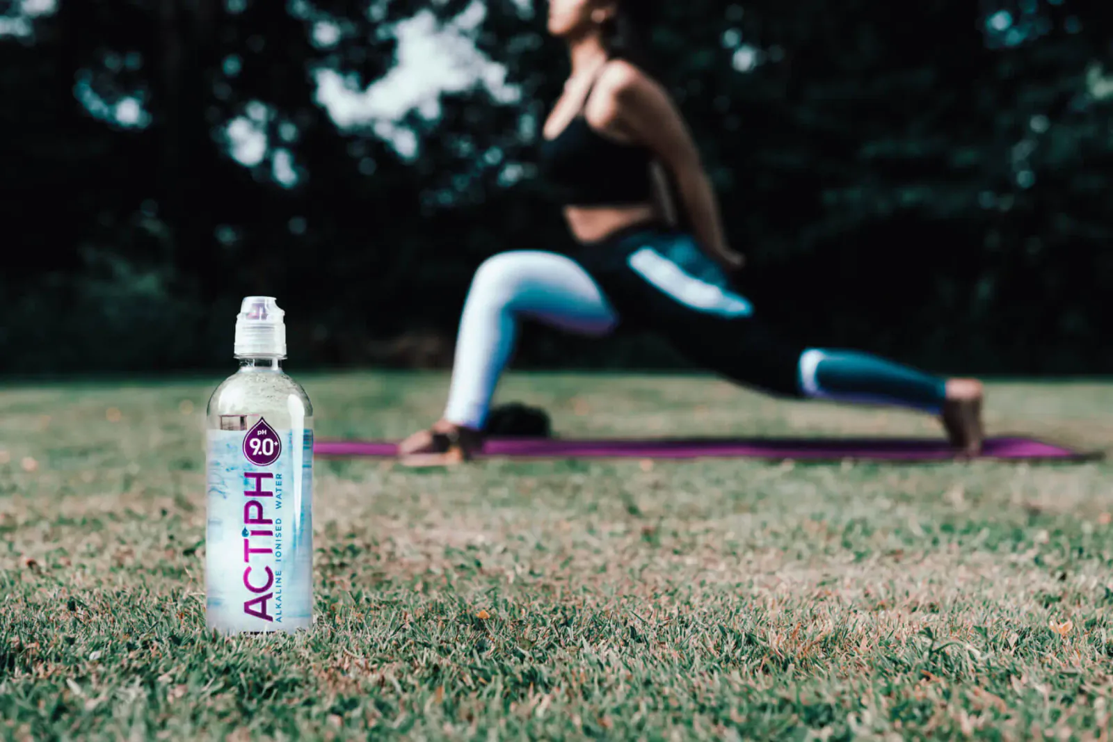 Actiph Alkaline Ionised Water bottle beside woman in warrior yoga pose on purple mat in grassy park