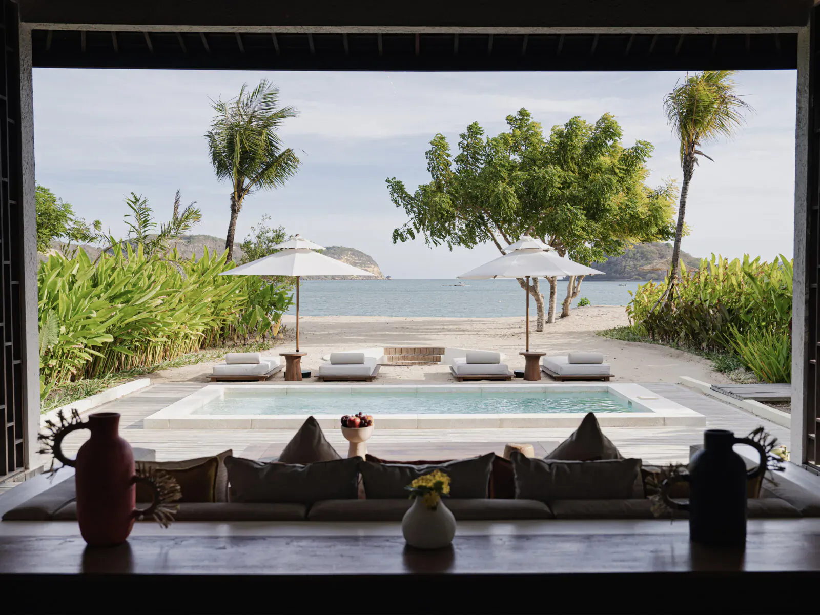 Kuara Lombok resort view from indoor seating: infinity pool, umbrellas, lush gardens, beach, and sea beyond palm trees.