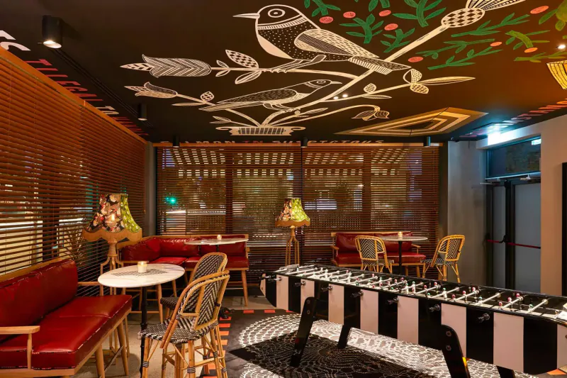 Stylish Mama London bar interior with bird mural ceiling, red seating, foosball table, lamps, and Halloween gallery vibe