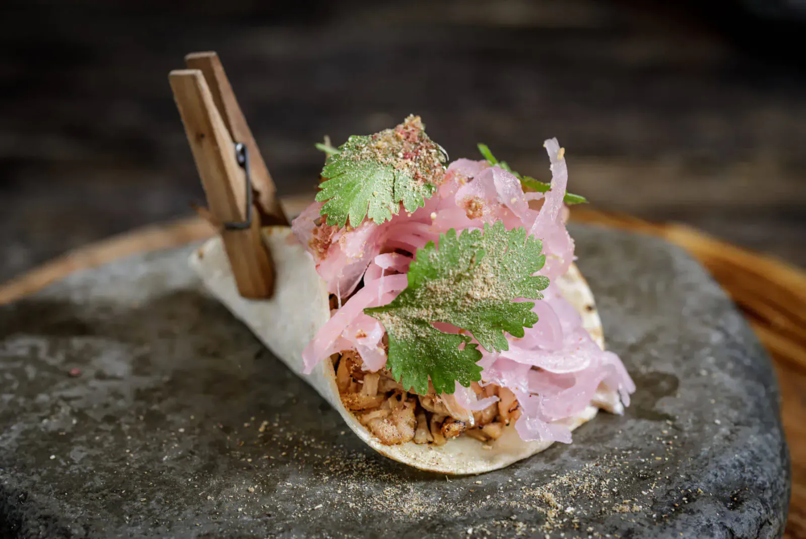 Close-up of a soft taco with pink pickled onions, cilantro, and shredded meat on a stone slab, held by wooden clips