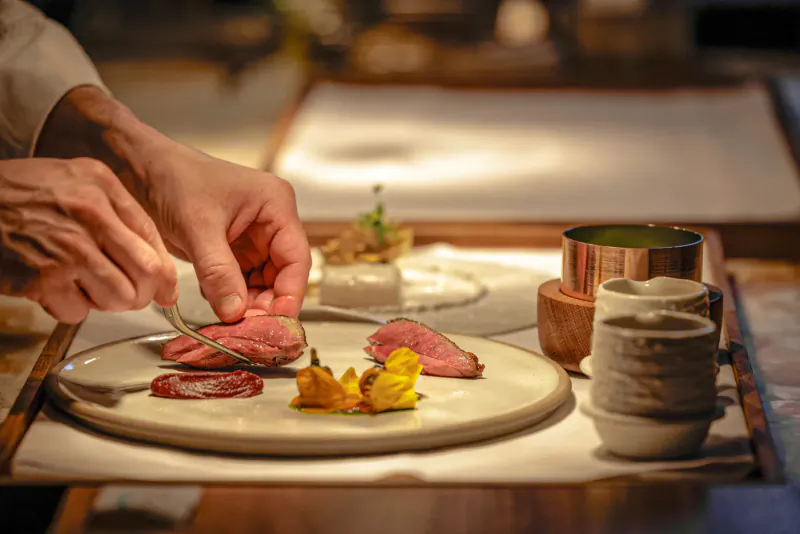 Chef's hands slicing pink duck breast on plate with garnishes, edible flowers, and ceramic cups at elegant restaurant table.