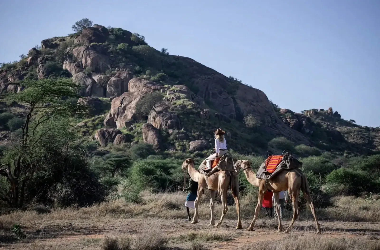 Woman rides camel led by another in Tumaren Camp safari, rocky green hills and acacia trees backdrop.