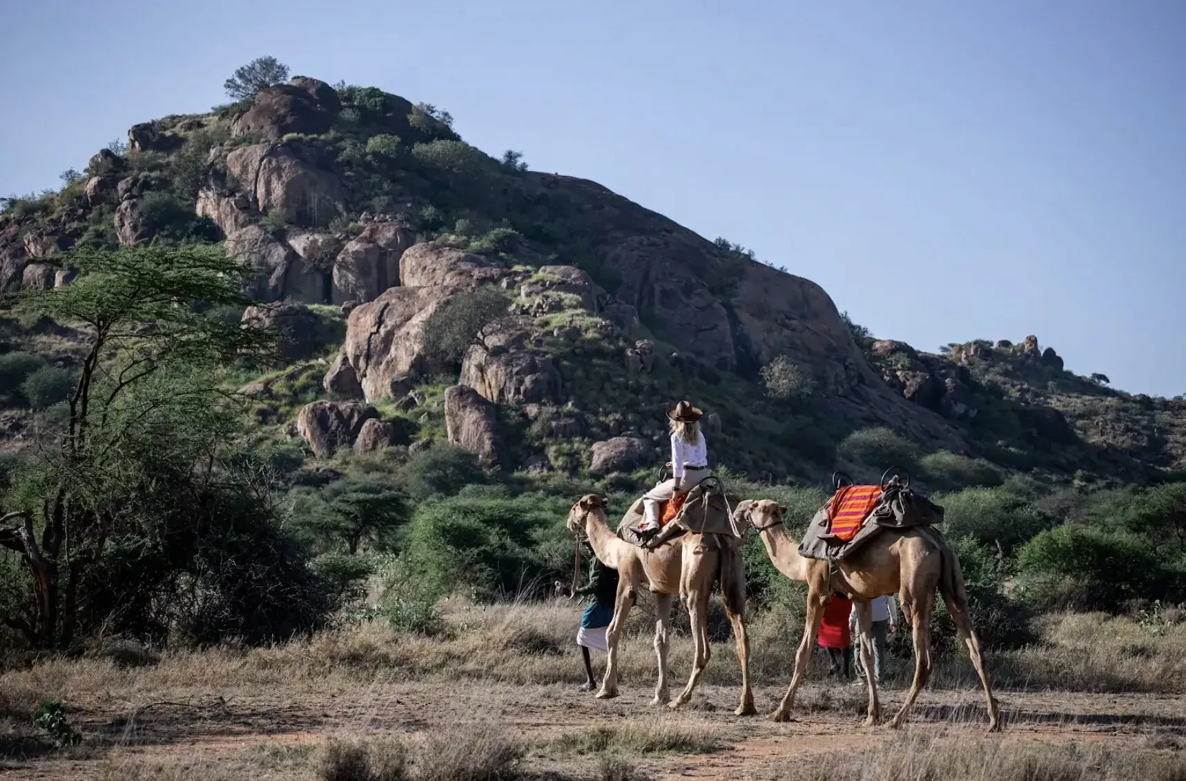 Woman rides camel led by another in Tumaren Camp safari, rocky green hills and acacia trees backdrop.