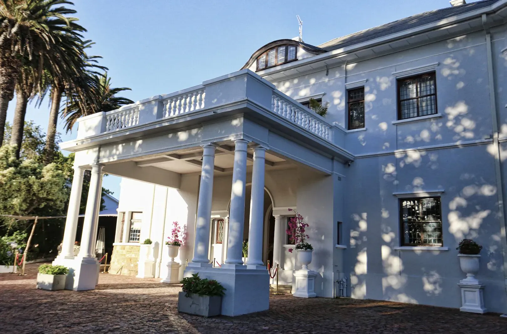 Elegant white colonial mansion with columns, balcony, palms, pink flowers, and potted plants at entrance under blue sky