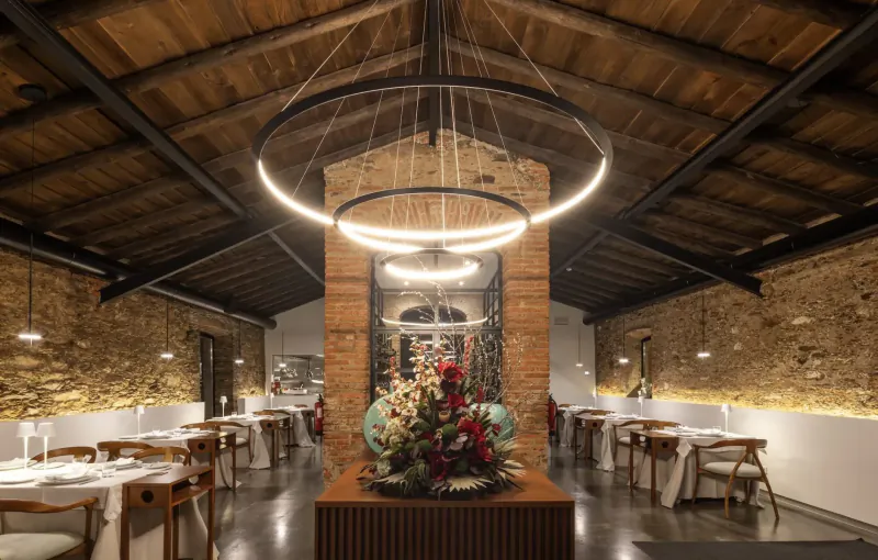 Elegant restaurant interior with wooden beams, large ring light fixture, brick walls, flower centerpiece, and white tables.