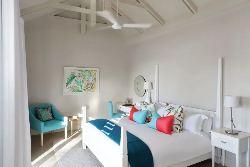 Bright bedroom in The Old Rectory with white four-poster bed, teal pillows, turquoise armchair, fan ceiling, tropical art.