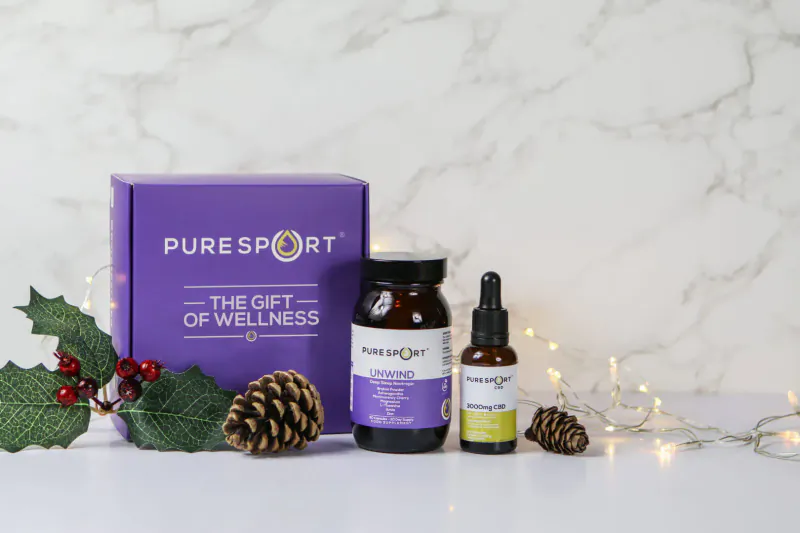 PureSport purple gift box 'The Gift of Wellness', amber jar, green dropper bottle on marble with holly, pinecones, fairy lights for Christmas.