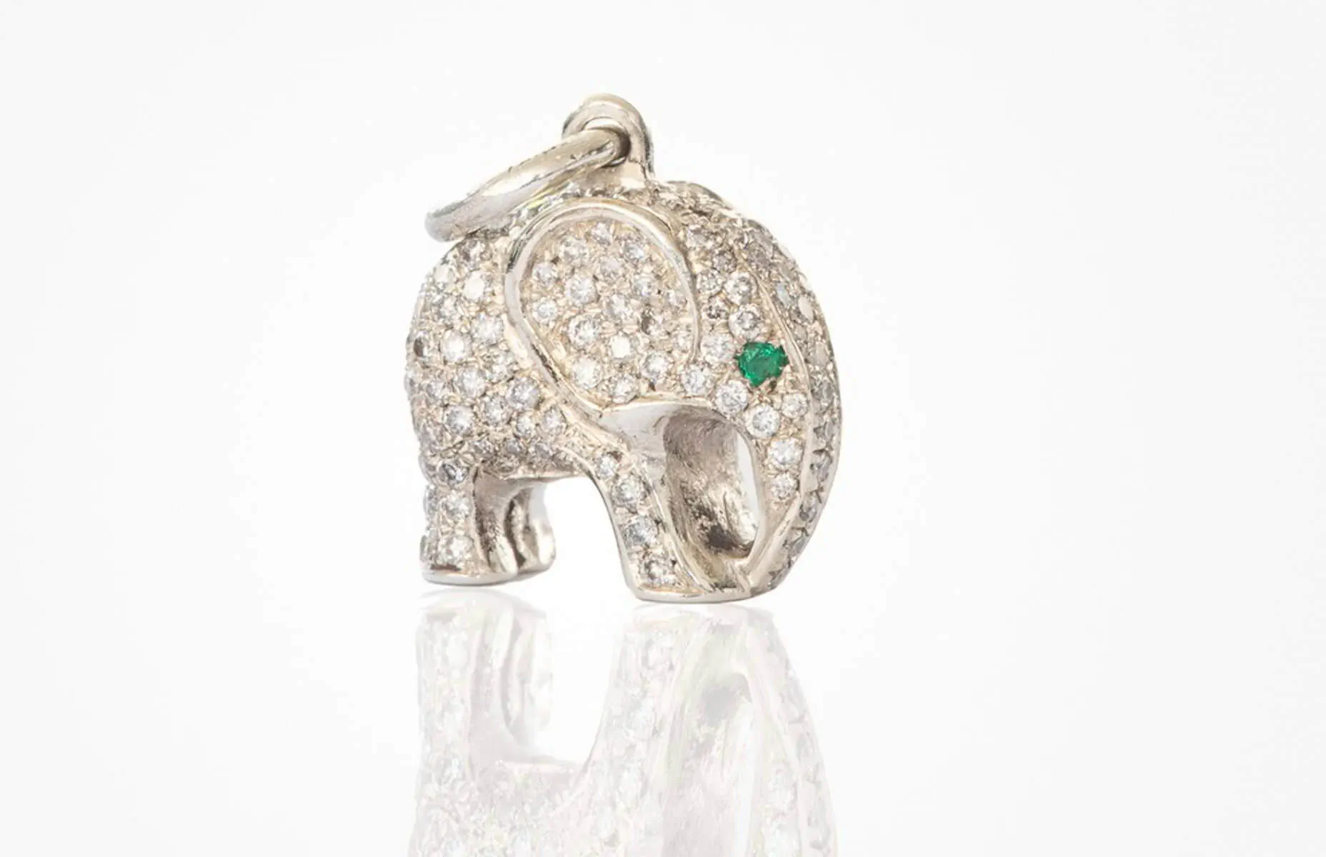 Silver diamond elephant pendant with green gem eye on chain, reflecting on white surface
