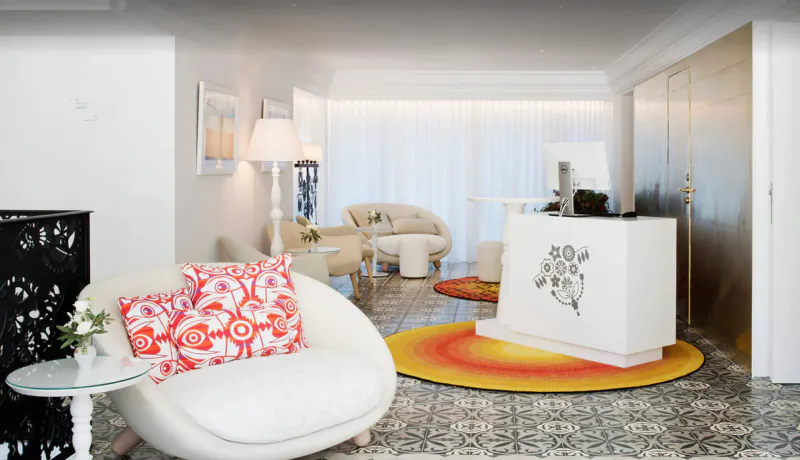 Marcel Wanders-designed hotel lobby interior with white curved sofa, red floral pillow, white reception desk with floral motif, orange rug, lamps, plants.