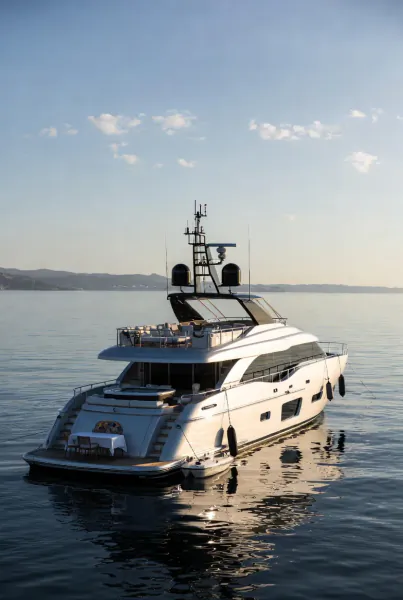 Sleek white luxury yacht viewed from rear, anchored on calm sea at sunset with distant mountains and clouds.