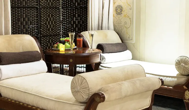 Elegant spa lounge at Bentley Hotel with two cream chaise lounges, bolsters, round table with champagne flutes, fruit, candle.