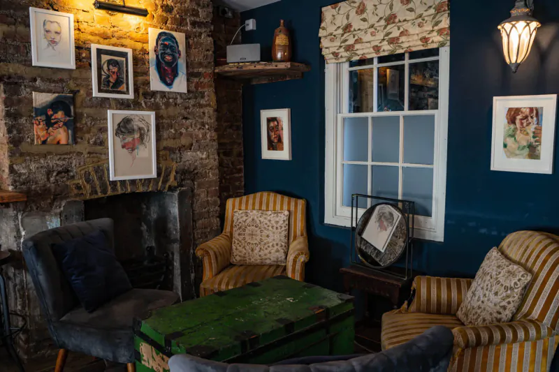 Cozy blue-walled room with exposed brick, vintage trunk table, patterned armchairs, and body-themed art portraits.