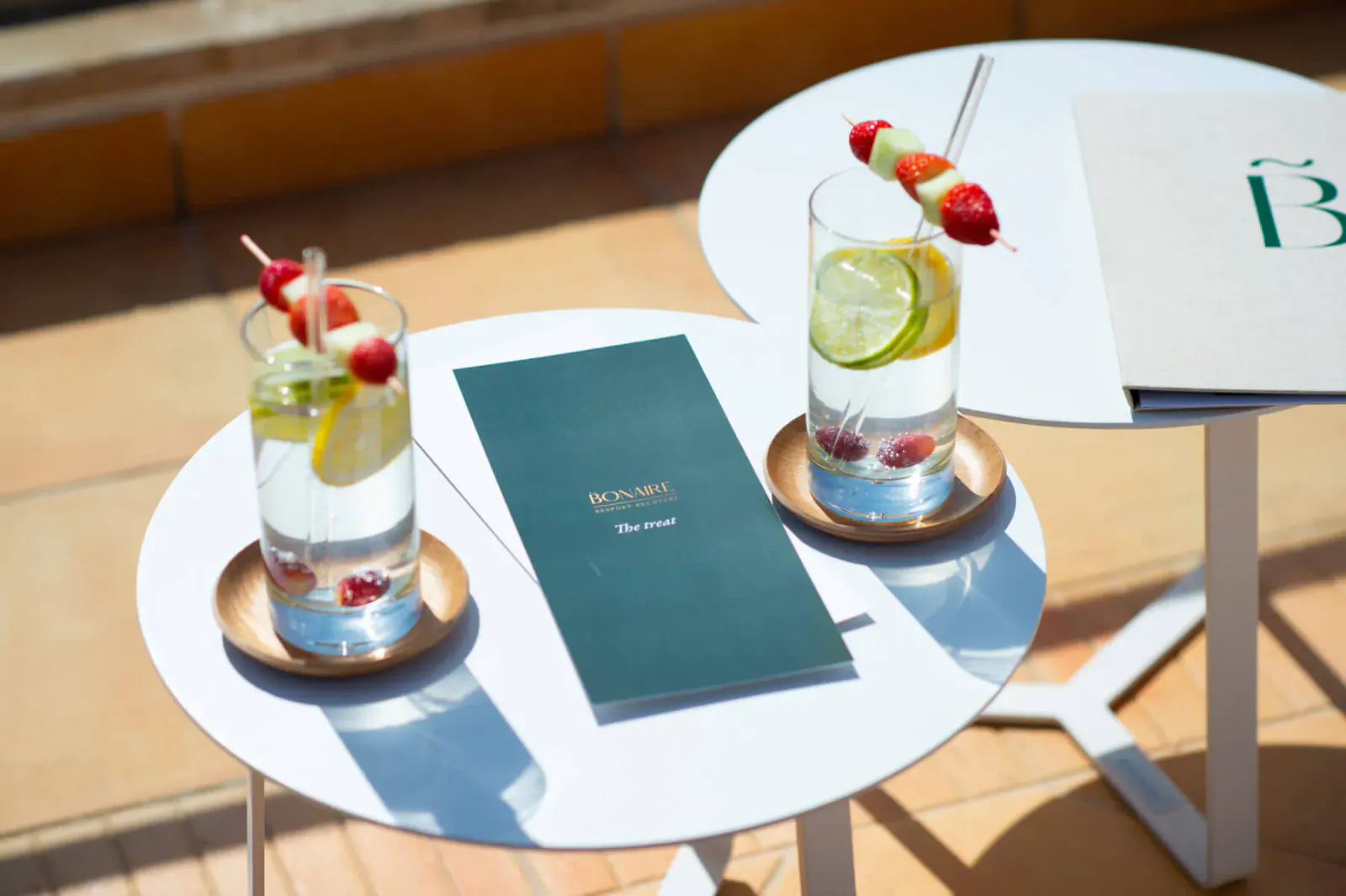 Two tall glasses of sparkling water with lime and raspberry skewers on gold saucers, beside Bonaire ReTREAT menus on white tables.