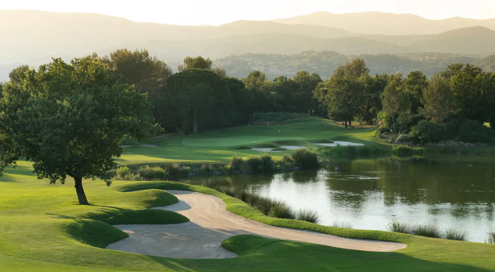 Sunset view of Terre Blanche golf course in Provence: lush fairway, sand trap, pond, trees, and distant mountains.