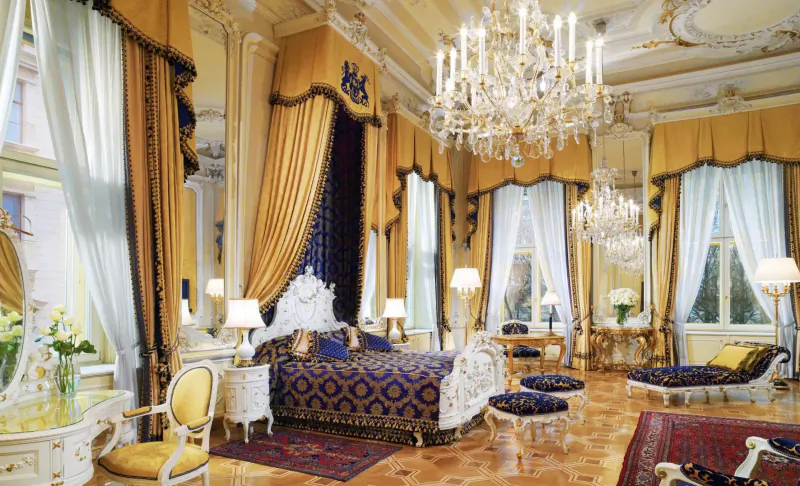 Opulent rococo bedroom in Hotel Imperial Vienna with ornate canopy bed, chandeliers, gold drapes, and parquet floor.