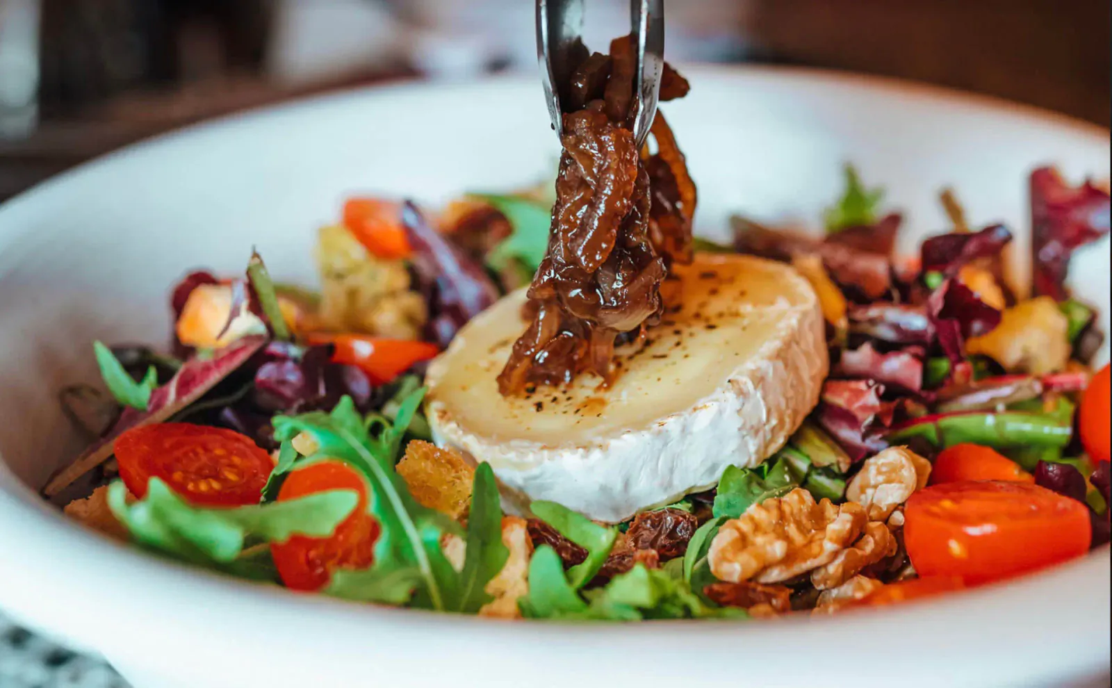 Close-up of vibrant salad in white bowl with baked brie cheese, pulled by tongs, tomatoes, walnuts, and greens