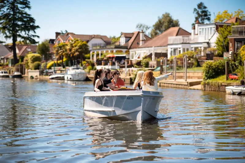 Group of four people in white boat on calm river, chatting amid riverside houses and trees under sunny sky.