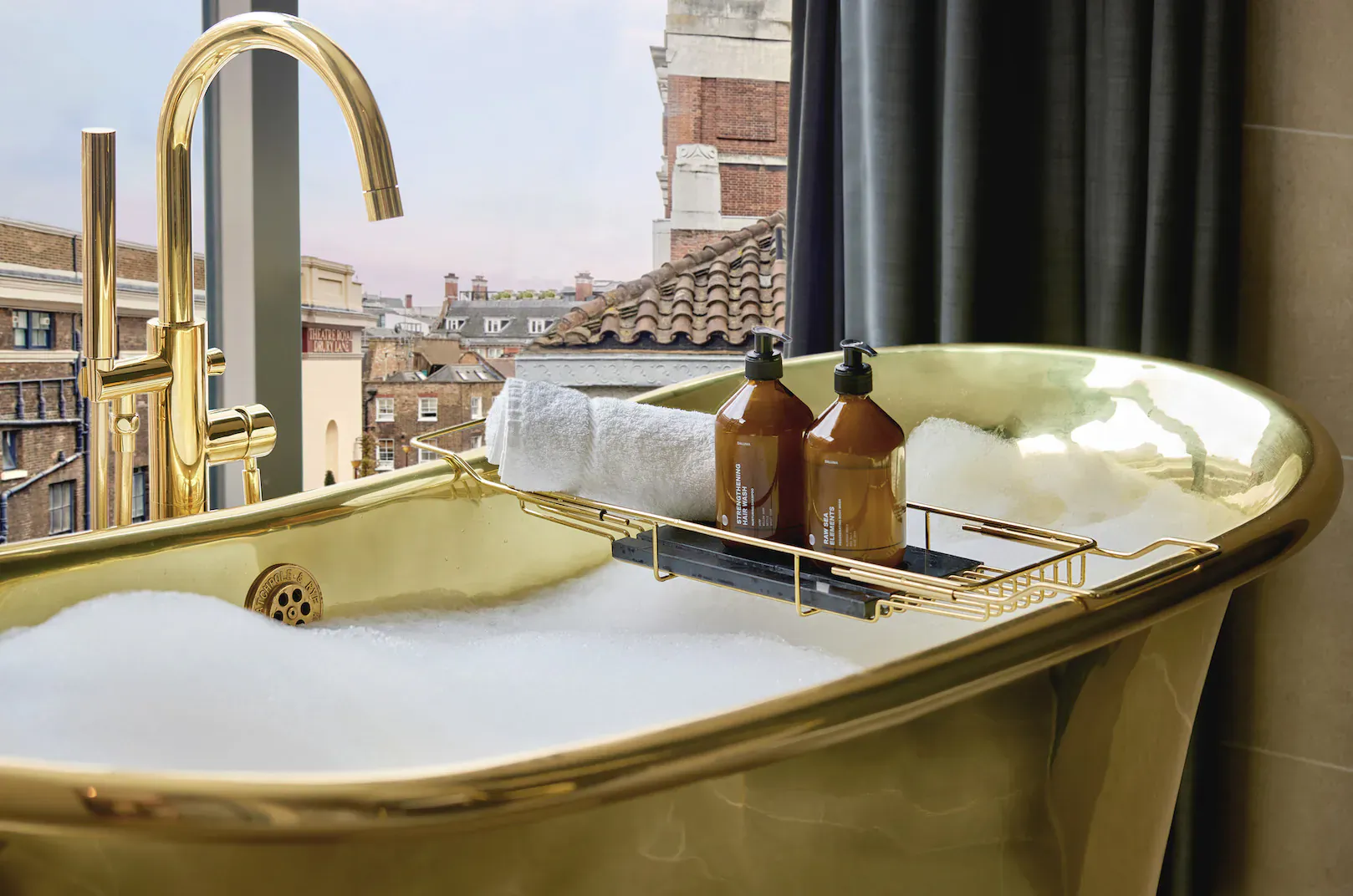 Luxurious gold bathtub filled with bubbles at Amano Hotel, with amber bottles, towel, brass faucet, and Covent Garden city view.