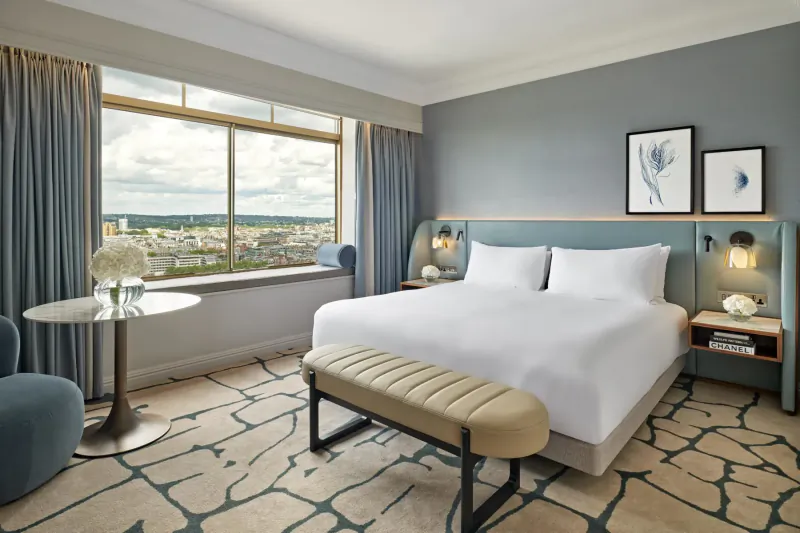 Elegant hotel room with king bed, blue headboard, bedside lamps, bench, side table with flowers, and city skyline view.