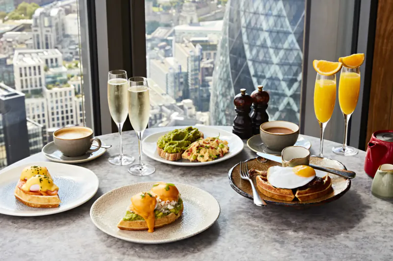 Duck & Waffle restaurant table with eggs Benedict, avocado toast, waffles, mimosas, coffee overlooking London skyline.