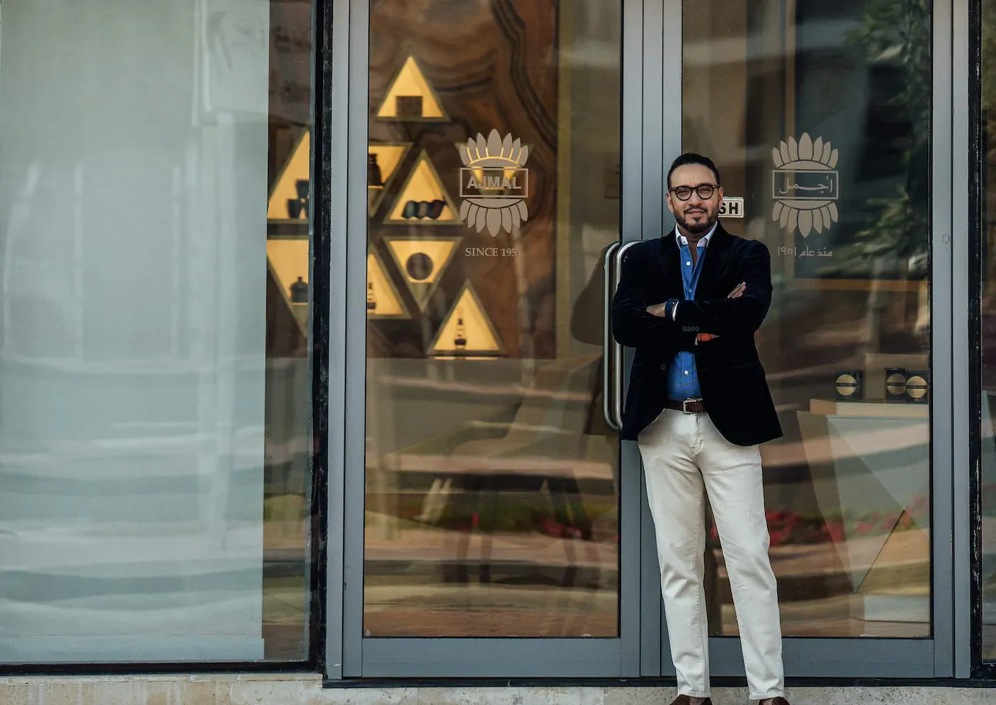 CEO Abdulla Ajmal in navy blazer and white pants, arms crossed, standing confidently outside Luxuria Lifestyle entrance with geometric logo.