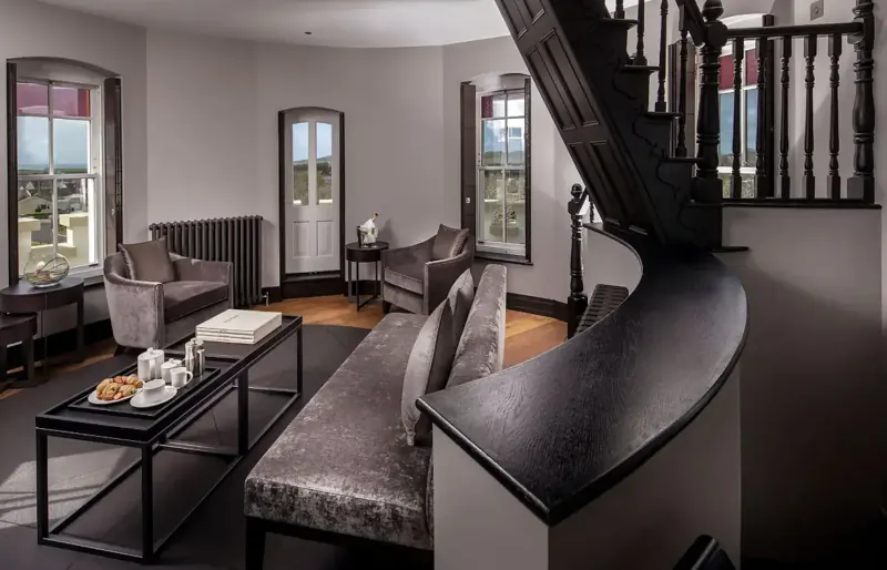 Elegant hotel lounge in Twr y Felin with grey velvet sofa, armchairs, tea service on coffee table, staircase, and window views.