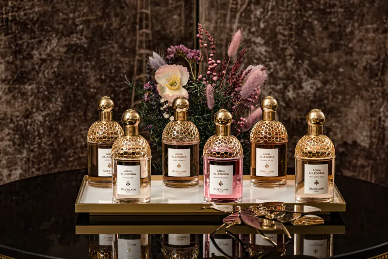 Six gold Guerlain perfume bottles on a black tray with pink flowers, against textured wall in elegant Parisian hotel setting