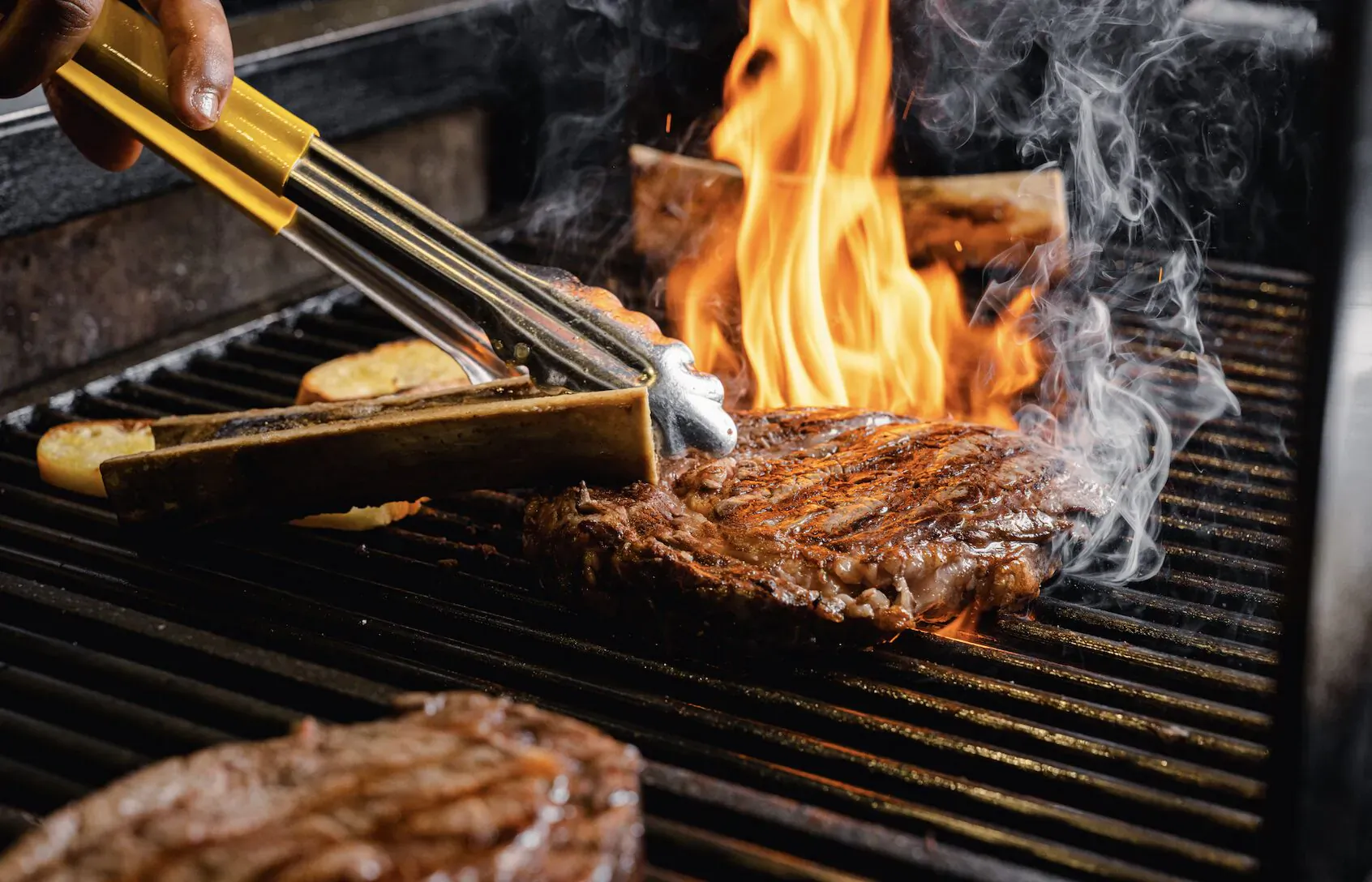 Hand using yellow tongs to flip seared steak over flames on black grill with potatoes and ribs