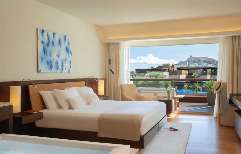 Luxurious hotel bedroom with king bed, balcony view of mountains, pool, and sea in Ibiza.