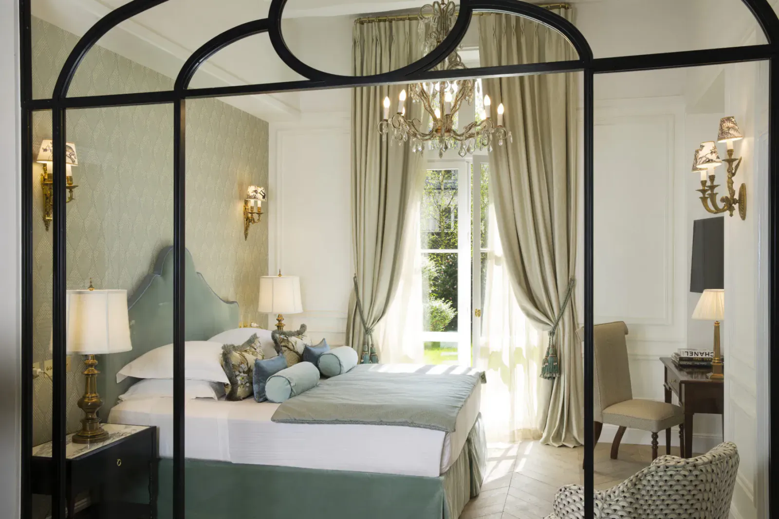 Elegant Parisian hotel bedroom with arched French doors, chandelier, tufted bed, and garden view at Relais Christine.