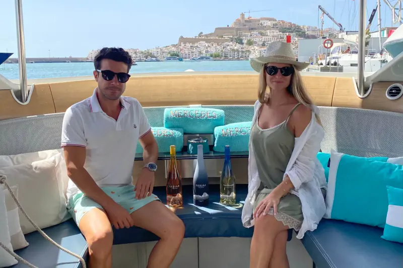 Smiling couple in sunglasses on luxury yacht, with Robiza rosé and white wine bottles, marina and castle view.