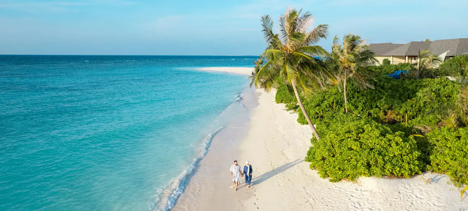 Aerial view of elderly couple walking hand-in-hand on white sand beach at Hideaway Beach Resort, turquoise ocean and palm trees.