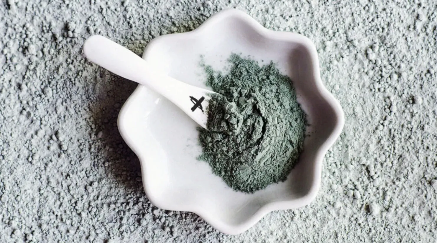 White scalloped bowl filled with green powder, white spoon with X mark resting in it on gray surface