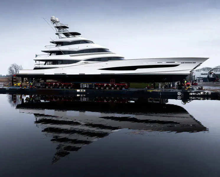 White multi-deck superyacht on a transporter in shipyard, reflected in water, overcast sky.