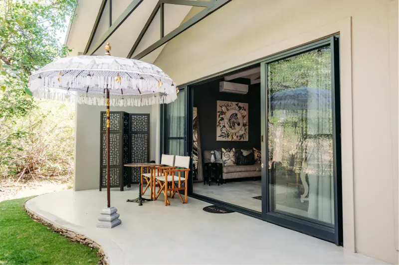 Luxury safari lodge patio with white Balinese umbrella, wooden table and chairs, open glass doors to modern lounge amid lush greenery.