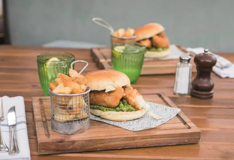 Fish burger with fries in metal basket, lime water glasses, salt and pepper on wooden board at Aztec Hotel table