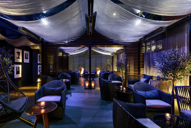 Elegant outdoor lounge at The May Fair Hotel with draped canopy, plush chairs, lanterns, and olive trees under night lights.