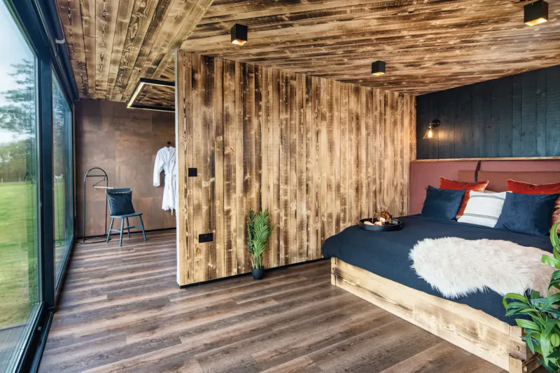 Cozy wooden bedroom in The Boathouse lodge with large window to green meadow, plush bed, robe on chair, plants.