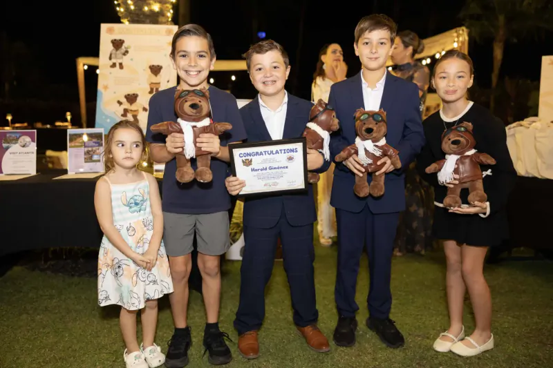 Smiling children in suits and dresses holding teddy bears and 'Make-A-Wish' certificate at outdoor event with backdrop.