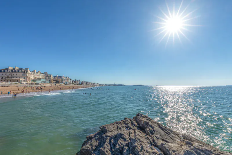 Sunny beach scene with sparkling sea, rocky foreground, sandy shore with people, and Grand Hotel des Thermes in background.