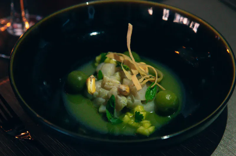 Close-up of ceviche in black bowl with green sauce, olives, herbs, crispy strips at Lio Ibiza restaurant