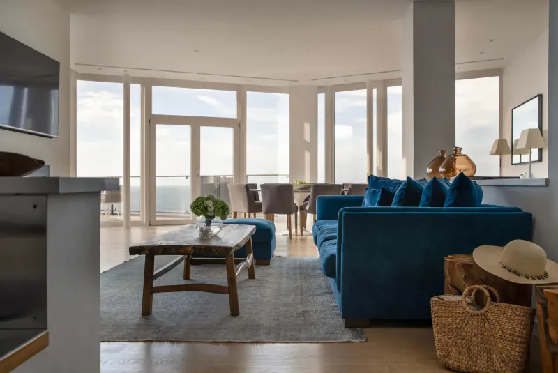 Spacious modern hotel suite with navy blue sofa, wood coffee table, plants, and panoramic sea views from large windows