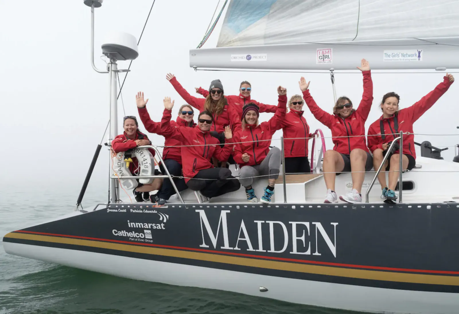 Group in red jackets waving on deck of yacht 'MAIDEN' with 'Ancasta' logo at sea