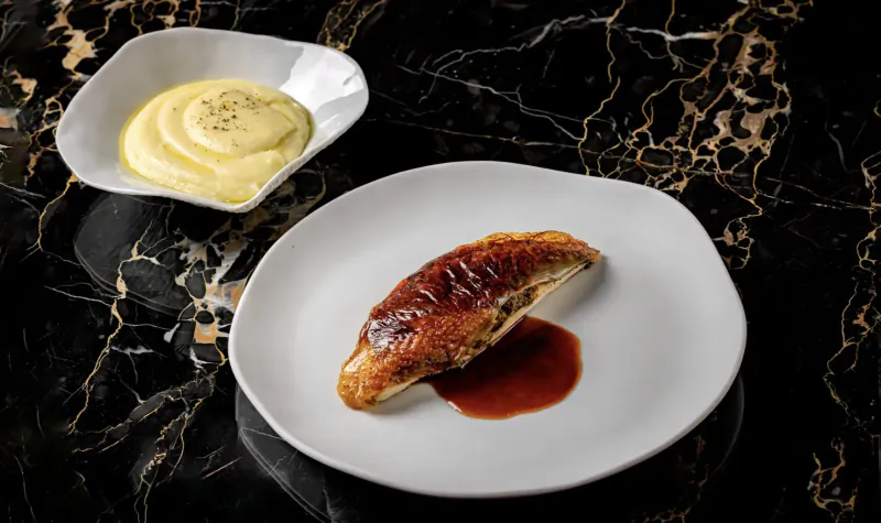 Grilled fish fillet with red sauce on white plate beside mashed potatoes with herbs, on black marble surface.