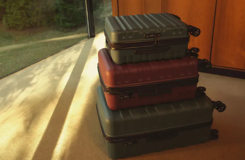 Stacked Antler Clifton suitcases in green and red winter hues by window with sunlight and greenery.
