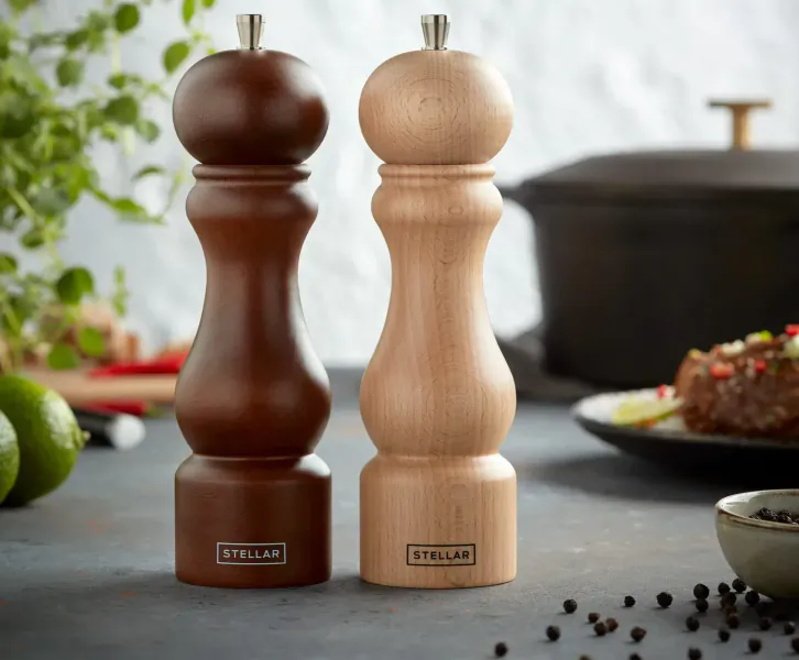 Stellar brown and light wood pepper mills on table with black pot, braised meat, limes, and peppers.