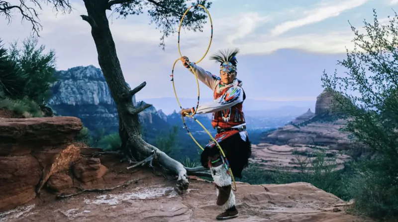 Native American dancer in feathered headdress twirling golden hoops on red rock cliff overlooking canyon at sunset