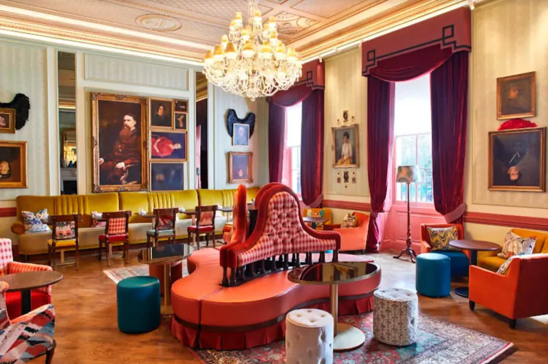 Opulent Home House gallery lounge in London with chandelier, large portraits, red velvet seating, and colorful chairs.